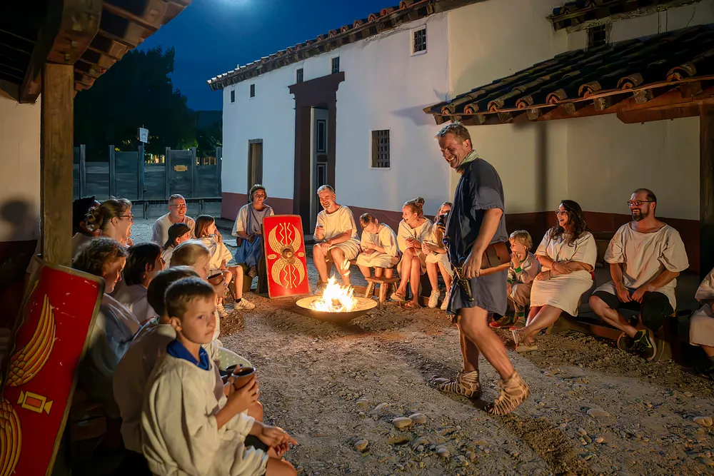Römisch gekleidete Männer und Frauen nachts an einem Lagerfeuer in historischer Kulisse