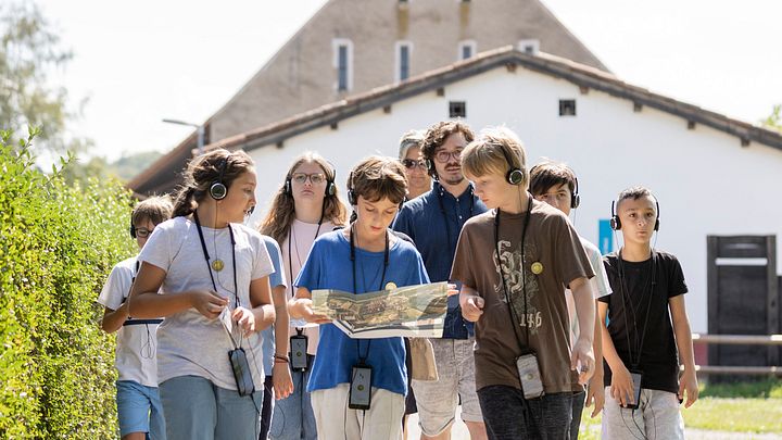 Eine Schulklasse unterwegs auf einem Kiesweg; im Hintergrund die rekonstruierte Unterkunft von Legionären.