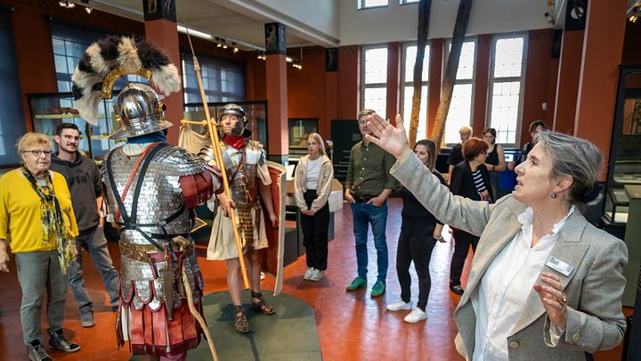 Eine Gruppe Besucherinnen und Besucher steht in der Ausstellung vom Vindonissa Museum (in der Mitte lebensgrosse Legionäre). Eine Museumsführerin steht davor und zeigt auf die beiden Männer.