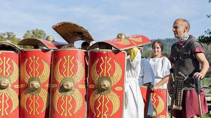 Eine Schulklasse in römischer Formation Schildkröte, rechts davon gibt ein Legionär Anweisungen.