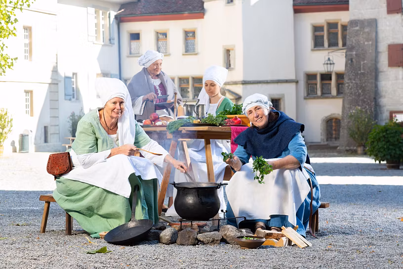 Mittelalterlich gekleidete Frauen sitzen im Hof von Schloss Lenzburg an einem Feuer und kochen. Im Hintergrund weitere Frauen an einem Tisch mit Essen aus dem Mittelalter