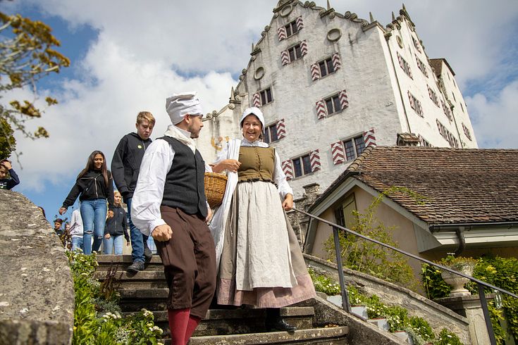 Eine Magd und ein Knecht vor Schloss Wildegg, dahinter eine Schulklasse.
