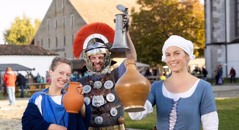 Eine Römerin, ein römischer Legionär sowie eine mittelalterliche Magd stehen nebeneinander. Sie prosten mit Wein direkt in die Kamera.