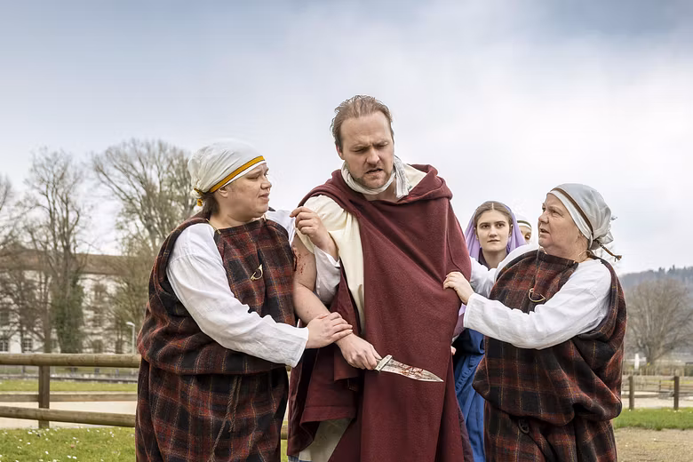 Mehrere Schauspieler in historischer Kleidung. Sie stehen nahe zusammen, der Mann in der Mitte hält einen Dolch in der Hand.