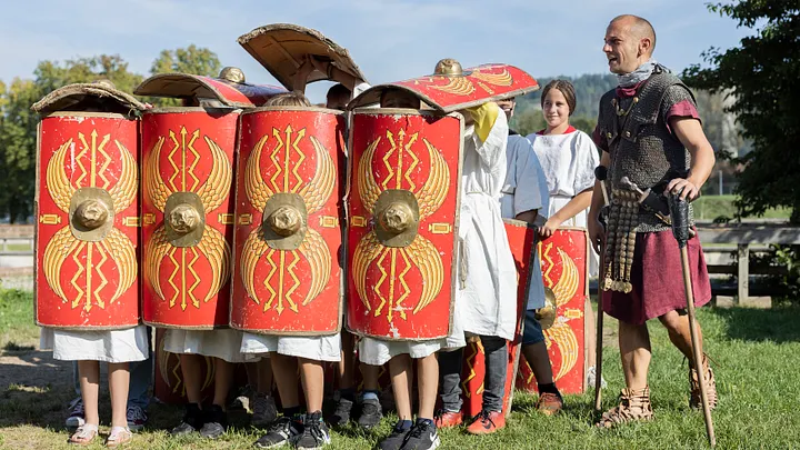 Eine Schulklasse in römischer Formation Schildkröte, rechts davon gibt ein Legionär Anweisungen.