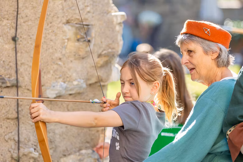 Ein Mädchen spannt auf Schloss Hallwyl einen Pfeilbogen auf. Daneben steht eine mittelalterlich gekleidete Frau.