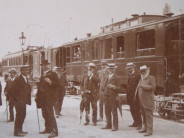 Schwarzweiss-Foto einer Männergruppe mit Anzügen und Hüten, die vor einem Zug mit mehreren Wagen stehen