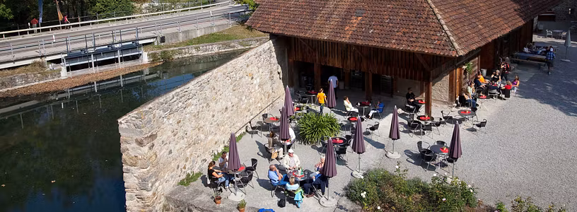 Blick von oben auf den Aussenbereich des Cafés von Schloss Hallwyl. An den Tischen im Schlosshof sitzen Besucherinnen und Besucher. Das Wetter ist sommerlich.