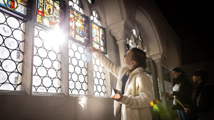 Eine Schülerin im Kreuzgang des Klosters Wettingen. Sie steht vor den Glasfenstern, durch die das Gegenlicht der Sonne in den Kreuzgang fällt.