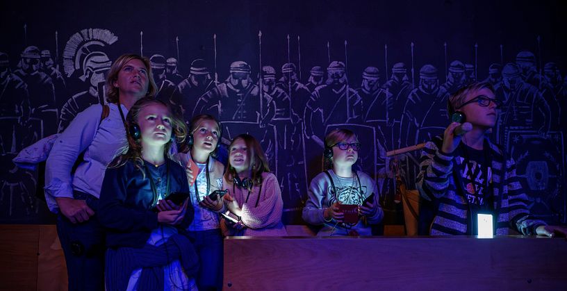 Eine Familie in einem dunklen Raum; die Displays der Audiogeräte um den Hals leuchten mystisch im Dunkeln. An der Rückwand Silhouetten von Legionären.