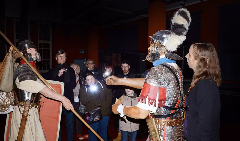 Besucherinnen und Besucher mit Taschenlampfen auf einer Führung im Vindonissa Museum. Die Gruppe steht vor zwei grossen Legionärsfiguren.
