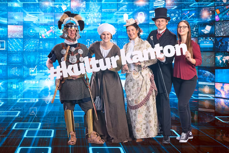 Vier historisch gekleidete Männer und Frauen (vom Römer bis zum Fabrikpatrons), rechts davon eine modern gekleidete Frau. Sie tragen den grossen Schriftzug "Kulturkanton" in ihren Händen.