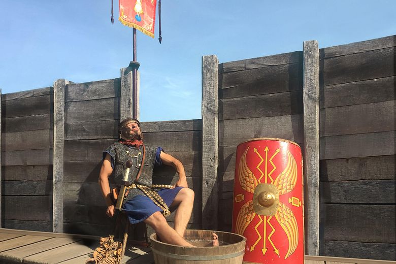 Ein Legionär sitzt auf einem Stuhl und blickt entspannt in die Sonne. Seine Füsse baden in einem Bottich mit Wasser.