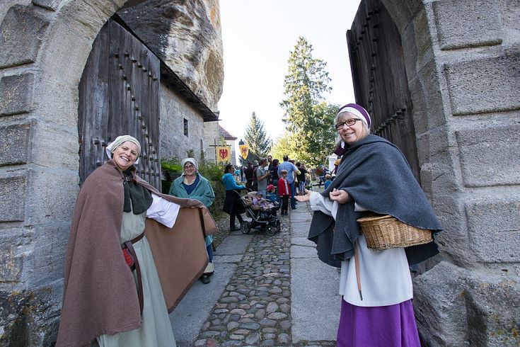 Zwei historisch gekleidete Freuen stehen vor dem Tor von Schloss Lenzburg und bitten Besucher mit einer einladenden Geste herein.