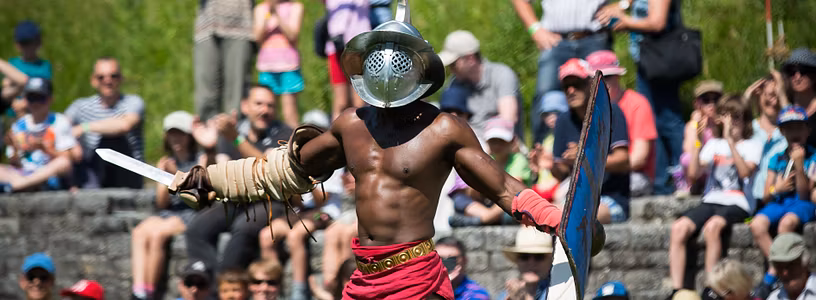 Ein kräftig gebauter Gladiator mit Schild und Schwert in Siegespose, im Hintergrund viel Publikum im Amphitheater Vindonissa (Veranstaltung Römertag).