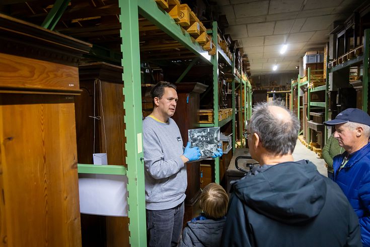 Ein Mann steht zwischen zwei Möbel im Sammlungszentrum Egliswil. Er hält ein schweiz-weiss-Bild in den Händen und spricht zu einer Besuchergruppe einer Führung.