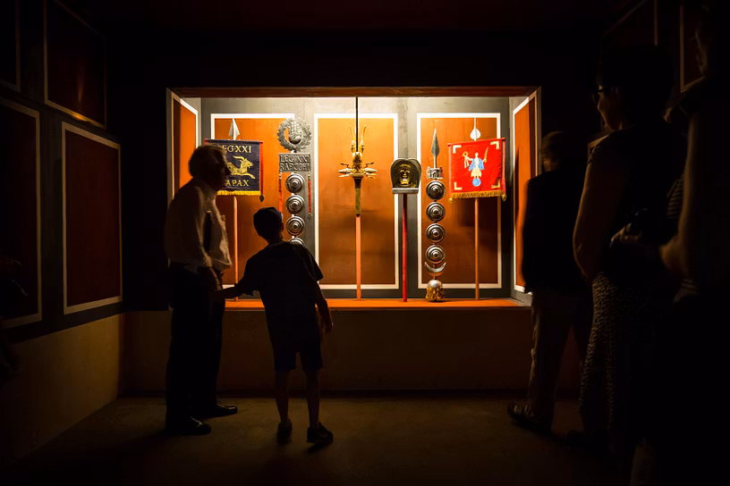 Mehrere Personen stehen im Ausstellungsraum "Fahnenheiligtum" im Legionärspfad Vindonissa und blicken in eine hell erleuchtete Vitrine mit Fahnen.