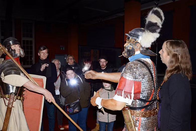 Besucherinnen und Besucher mit Taschenlampfen auf einer Führung im Vindonissa Museum. Die Gruppe steht vor zwei grossen Legionärsfiguren.