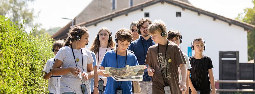 Eine Schulklasse unterwegs auf einem Kiesweg; im Hintergrund die rekonstruierte Unterkunft von Legionären.