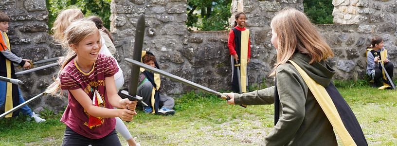 Workshop "Feste Burg Schloss Hallwyl" Kinder haben zusammen spass im Schwertkampf mit Zinnen im Hintergrund