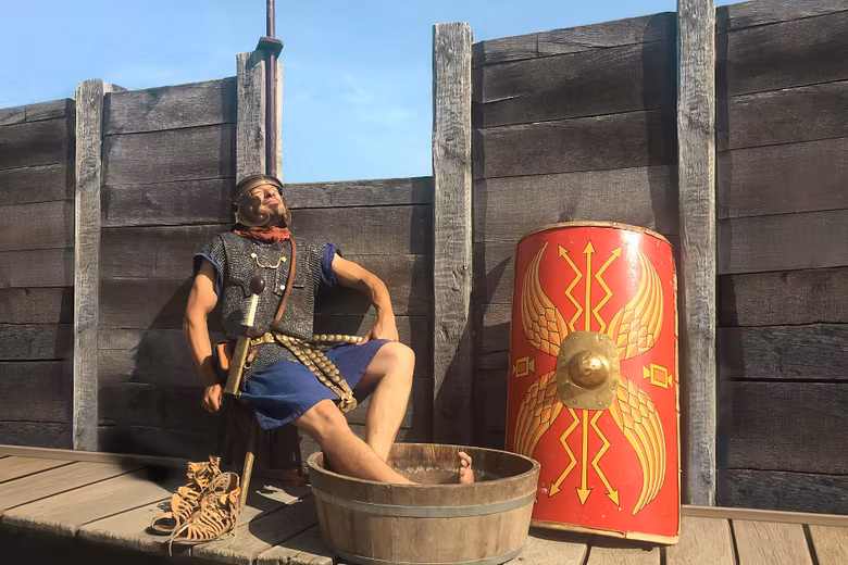 Ein römischer Legionär sitzt auf einem Stuhl und blickt entspannt in die Sonne. Seine Füsse liegen in einem Bottich mit Wasser.
