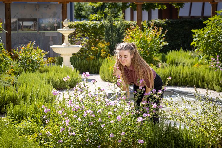 Römischer Garten Vindonissa Museum: Eine junge Frau steht inmitten vieler grüner Pflanzen und riecht an einer Blüte. Im Hintergrund ein antiker Brunnen.