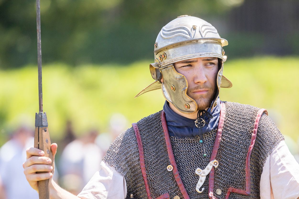 Auf dem Bild ist der obere Körperteil eines Legionärs zu sehen. Er trägt einen Helm und ein Kettenhemd, das seine Schultern und Brust schützt. In der linken Hand hält er einen Wurfspeer. Der Hintergrund des Bildes ist von einem verschwommenen Grün umgeben.