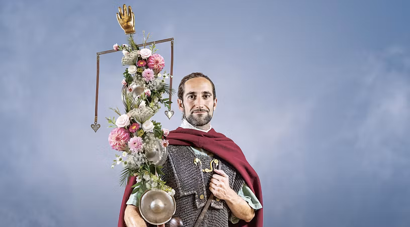 Ein römischer Legionär mit einer Standarte, die mit Blumen geschmückt ist. Er blickt direkt in die Kamera. Der Hintergrund ist blau (mit Wolken)