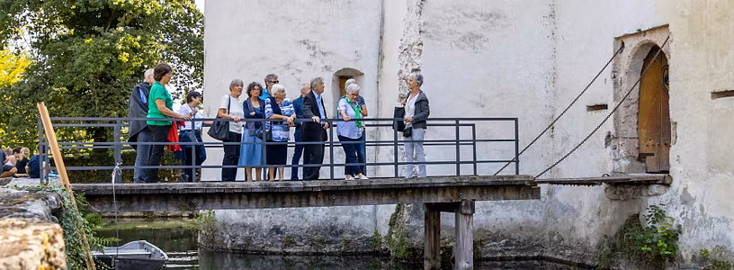 Führung auf Schloss Hallwyl: Eine Besuchergruppe steht auf einer Brücke zwischen Hof und dem Eingang zu Schloss Hallwyl, unter der Brücke Wasser des Schlossgrabens und ein Boot. 