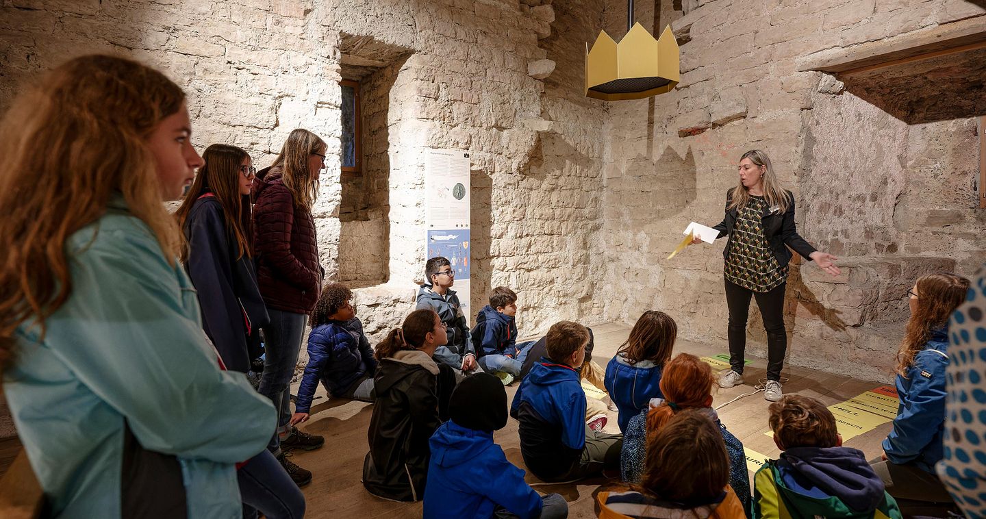Eine Schulklasse sitzt in einem Raum von Schloss Habsburg und hört einer Lehrerin zu, die etwas erklärt. 