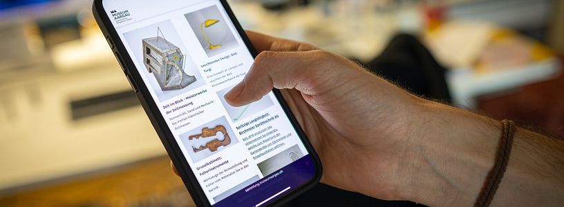Grosses Handy in der Hand eines Benutzers, auf dem Bildschirm ist die "Sammlung online" von Museum Aargau dargestellt.