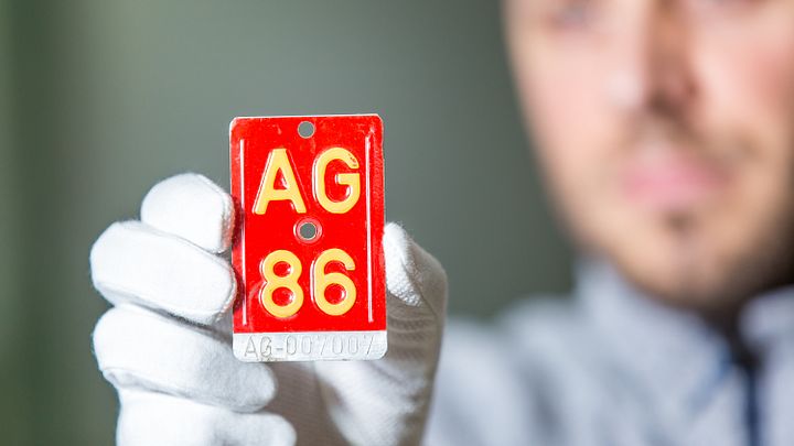 Rote Velonummer mit der Aufschrift "AG 86" in der Hand eines Mannes mit weissen Handschuhen. Im Hintergrund in Unschärfe der Kopf des Mannes (er hält die Velonummer in die Kamera).