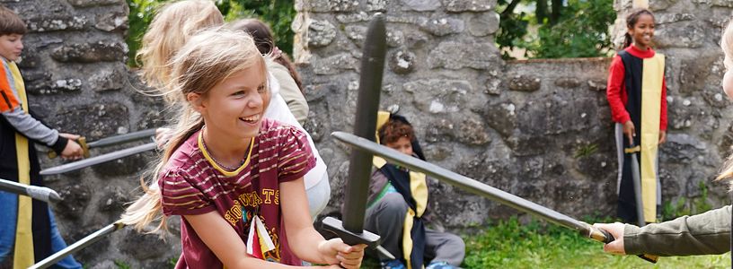 Workshop "Feste Burg Schloss Hallwyl" Kinder haben zusammen spass im Schwertkampf mit Zinnen im Hintergrund
