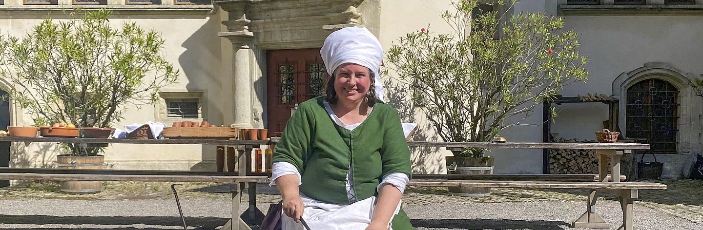 Eine mittelalterlich gekleidete Frau sitzt an einem Feuer im Hof von Schloss Lenzburg und kocht. 