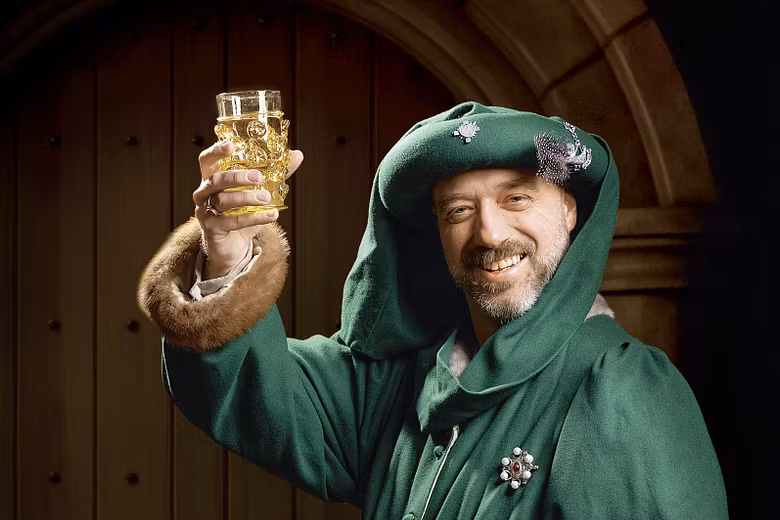 Ein mittelalterlich gekleideter Mann. Er blickt direkt in die Kamera und hält feiernd ein Glas mit Bier in die Höhe.