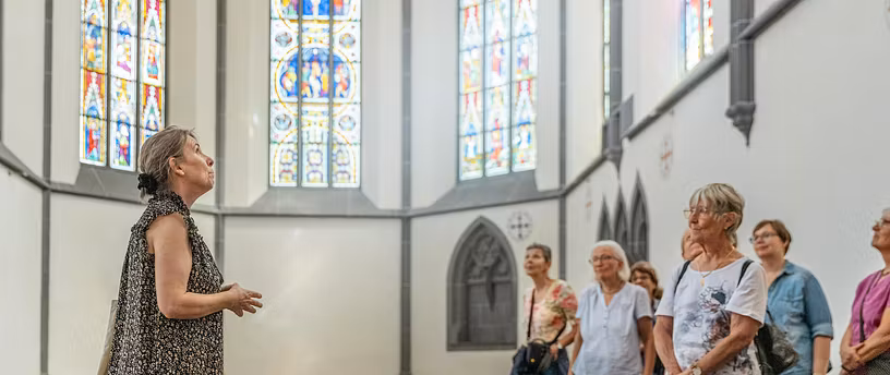 Eine Museumsführerin mit einer Gästegruppe im Chor der Klosterkirche Königsfelden; im Hintergrund die berühmten Glasfenster.
