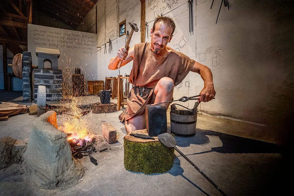 Ein römischer Schmied in der Schmiede im Legionärspfad Vindonissa. Er schlägt mit einem Hammer auf den Amboss und blickt direkt in die Kamera. Links ein Feuer mit etwas Glut.
