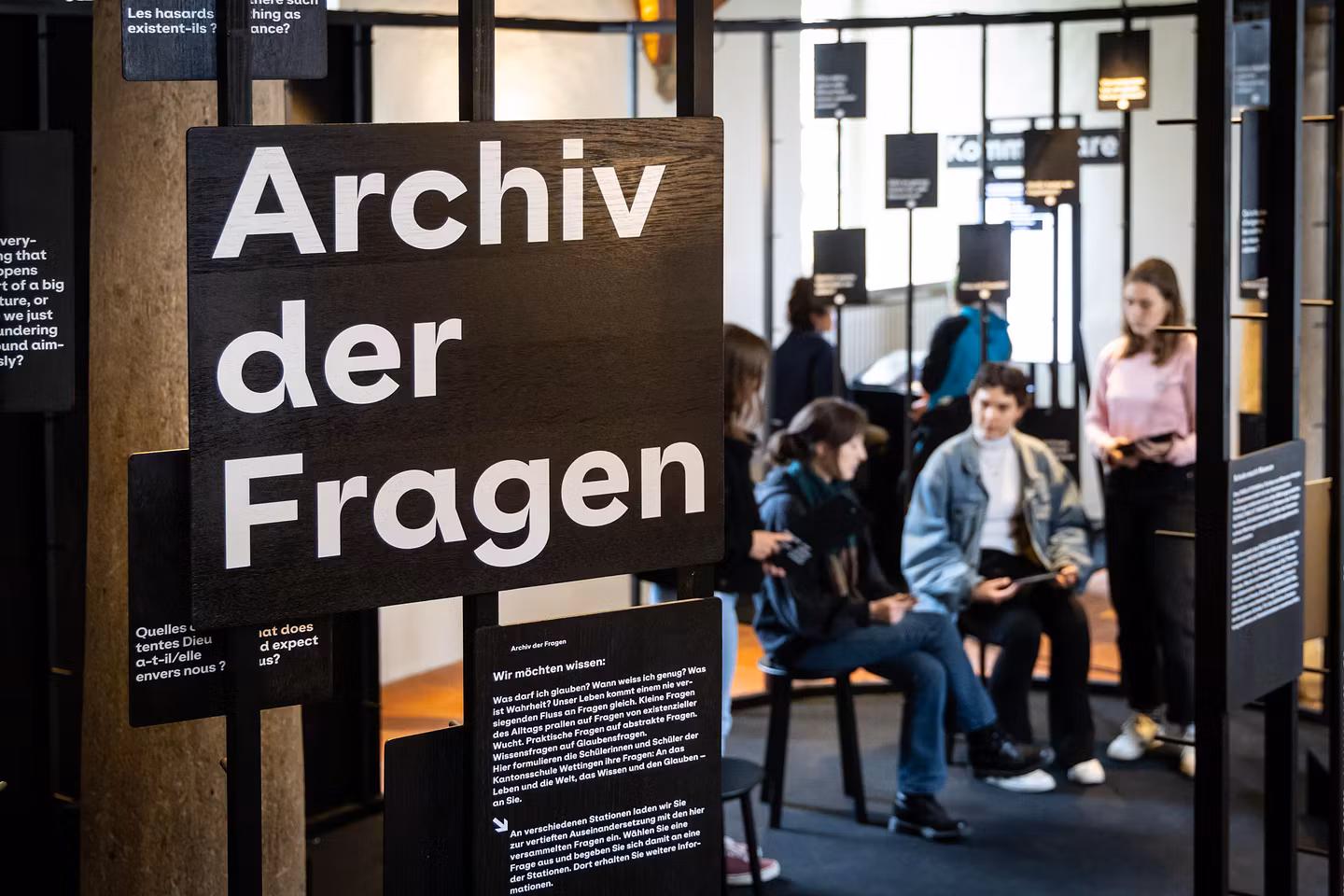 Ausstellungsraum im Parlatorium der Klosterhalbinsel Wettingen: Ein grosses Schild mit dem Text "Archiv der Fragen", im Hintergrund sind mehrere Personen zu sehen, die diskutieren.