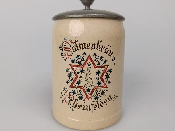 Bierhumpen mit Gravur und Deckel vor einem weissen Hintergrund. Die Gravur zeigt einen roten sechszackigen Stern, der von Pflanzen und Blättern umschlungen ist.
