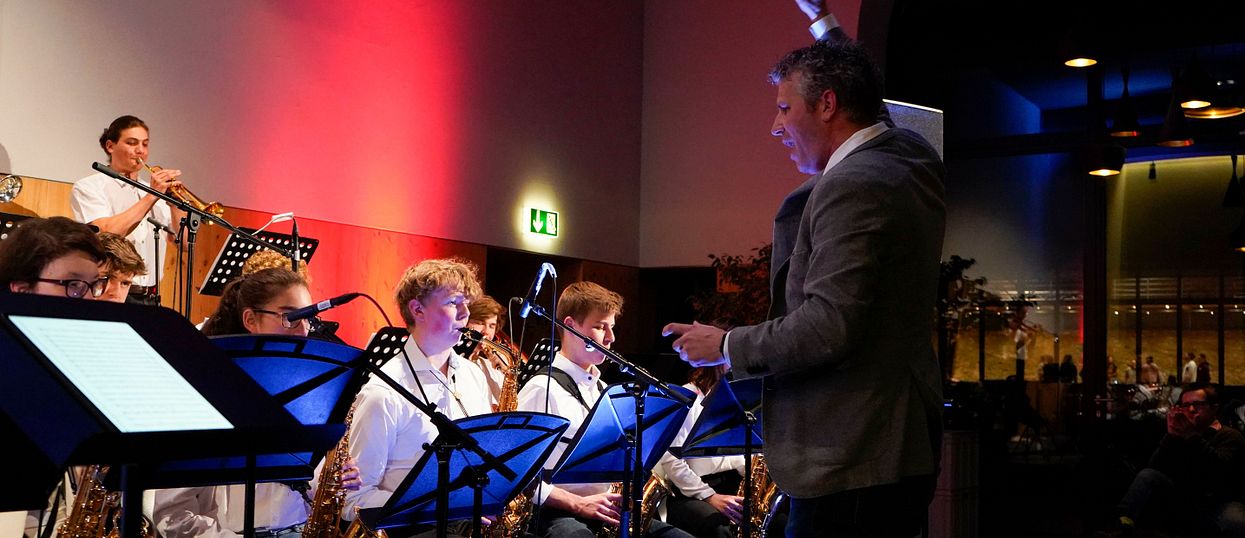 Im Bild sind links mehrere musizierende Personen zu sehen. Alle spielen verschiedene Blasinstrumente und haben ein weisses Hemd an. Vor den sitzenden Bandmitglieder steht der Dirigent in einem grauen Jackett.  