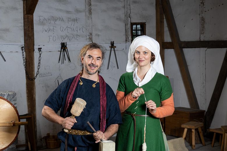 Ein römischer Steinmetz und eine mittelalterlich gekleidete Frau mit Wolle. Beide stehen in einem Werkraum und blicken direkt in die Kamera.