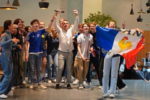Eine Gruppe Schülerinnen und Schüler steht mit einer Lehrperson auf einer Bühne und singt. Eine Schülerin hält eine französische Flagge hoch.