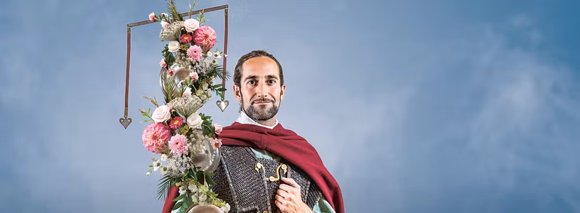 Ein römischer Legionär mit Standarte (geschmückt mit Blumen) vor blauem Hintergrund.