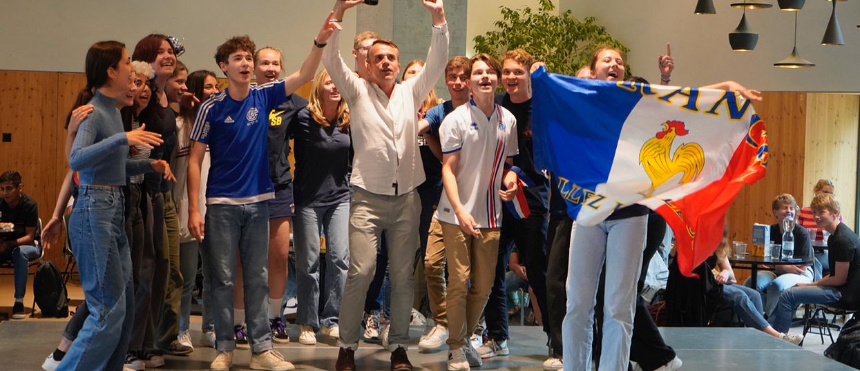 Eine Gruppe Schülerinnen und Schüler steht mit einer Lehrperson auf einer Bühne und singt. Eine Schülerin hält eine französische Flagge hoch.