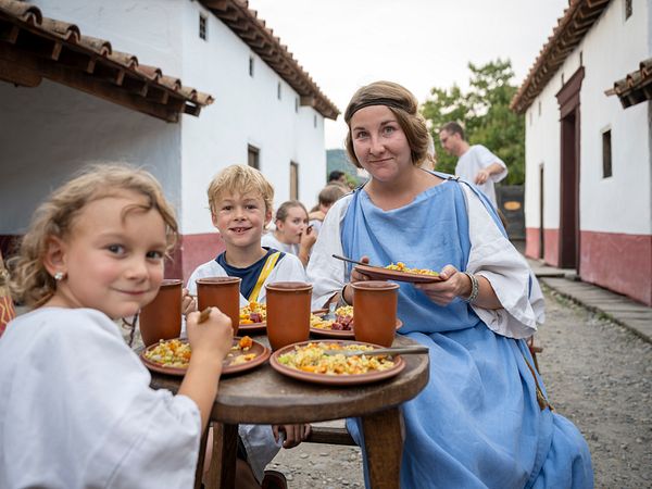 Eine in blauem Gewand gekleidete Römerin und zwei Kinder essen an einem Tisch. Sie blicken direkt in die Kamera und lachen.