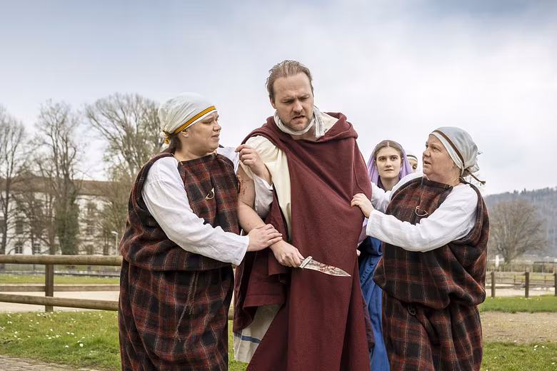 Mehrere Schauspieler in historischer Kleidung. Sie stehen nahe zusammen, der Mann in der Mitte hält einen Dolch in der Hand.