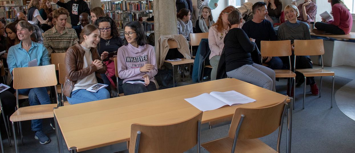 Ganz vorne ist ein Tisch mit zwei Stühlen gegen ein Publikum gerichtet, das aus mehreren Jugendlichen und Erwachsenen besteht. Gewisse Personen stehen im Hintergrund noch. Dahinter sind Bücherregale zu sehen, es handelt sich um eine Bibliothek. In der Mitte des Raumes gibt es vier Steinsäulen, die sich von vorne bis hinten ziehen.