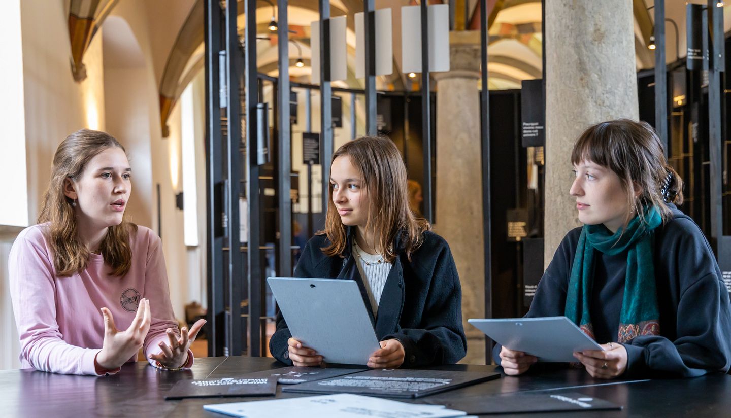 Ausstellung "Parlatorium auf der Klosterhalbinsel Wettingen": Drei Schülerinnen sitzen an einem Tisch und diskutieren angeregt über Fragen, die auf grossen Tafeln vor ihnen stehen.