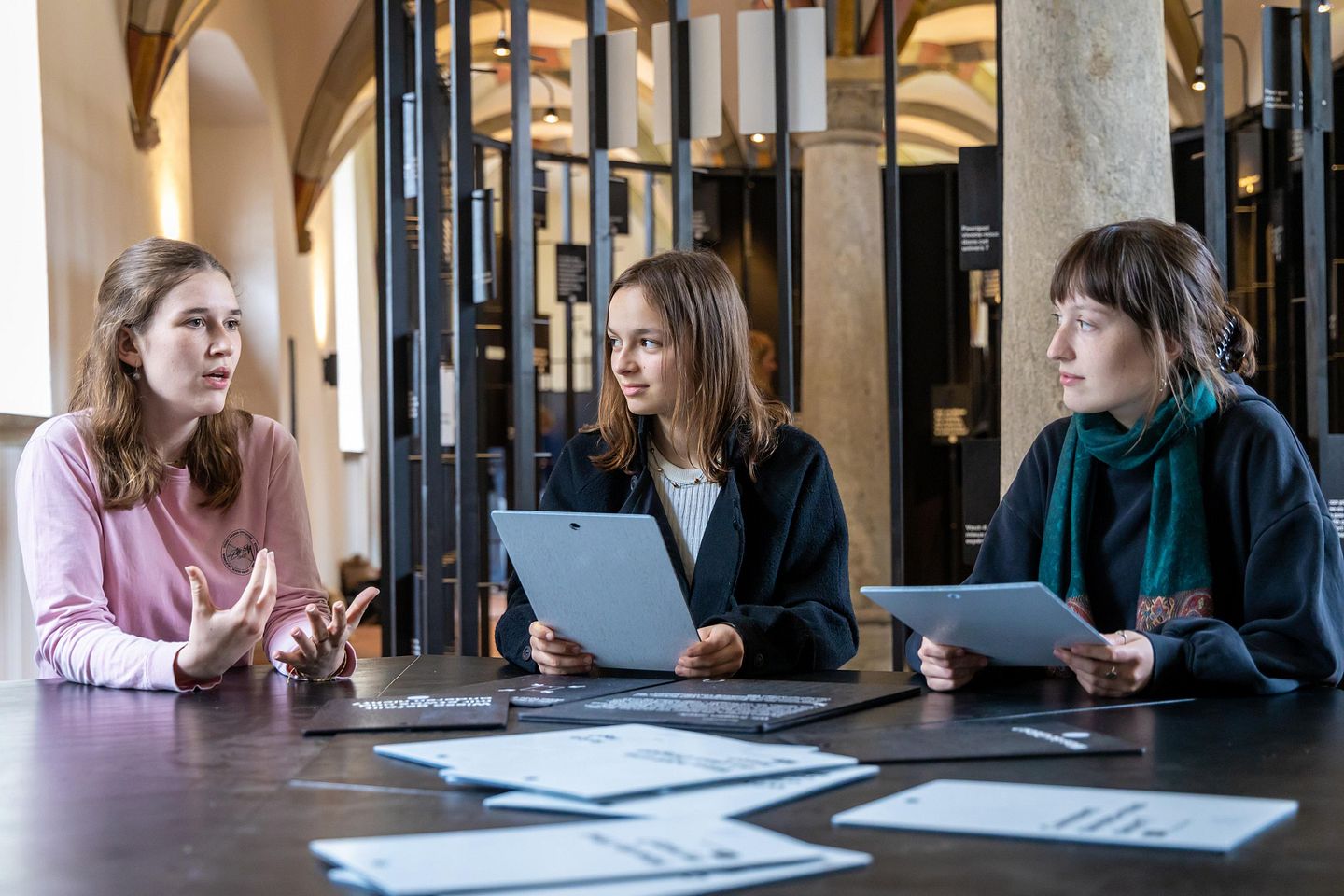 Ausstellungsraum im Parlatorium der Klosterhalbinsel Wettingen: Drei Schülerinnen sitzen mit Unterlagen am Tisch und diskutieren miteinander.
