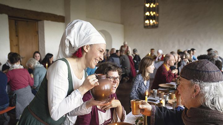 Gäste sitzen im kleinen Rittersaal von Schloss Lenzburg an langen Tischen und essen gemeinsam. Eine Frau in mittelalterlicher Kleidung giesst Getränke nach.